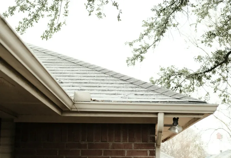 A brick house seen from below at the eaves, with the gutter running along the roof edge and the branches of a mature tree overhanging it.