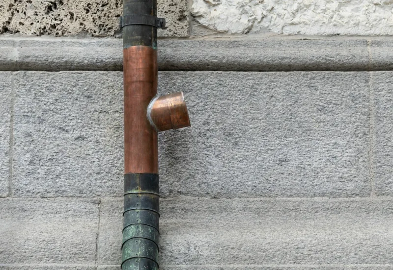 A copper downpipe running down a grey stone wall and ending in a copper shoe that throws the water clear of the base.