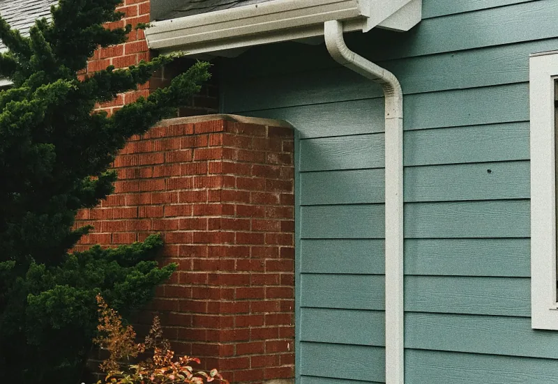 A white gutter running along the eaves of a house clad in green clapboard, turning down into a downspout beside a brick chimney.