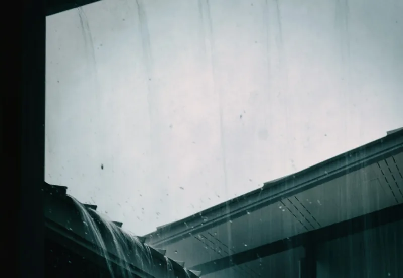 Rain sheeting off the edge of a pale roof and falling past the gutter beneath it.